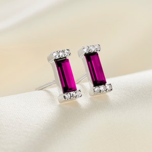 May include: A pair of silver earrings with a rectangular, dark purple gemstone set in each earring. The earrings are surrounded by small, clear, round gemstones.