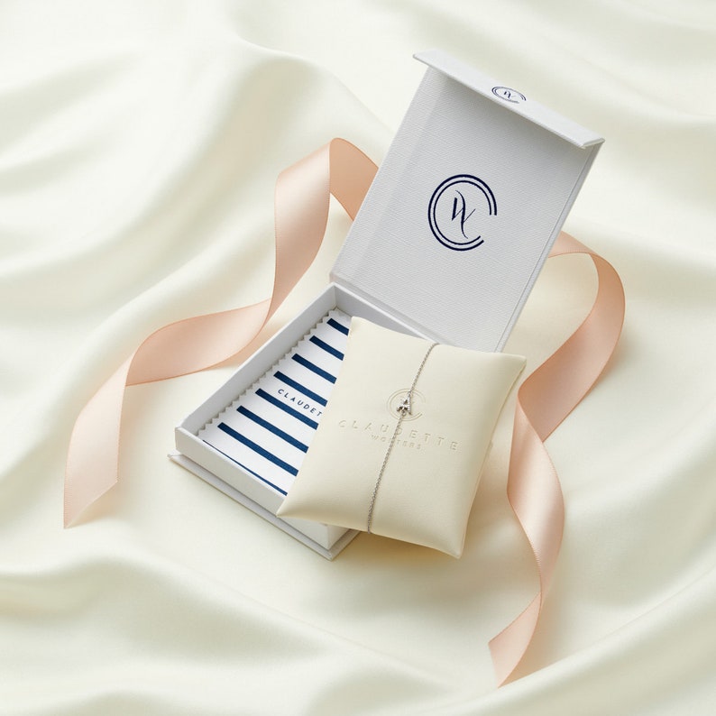 May include: A white box with a blue and white striped interior and a white pouch with a silver chain necklace inside. The box has a logo that says "Claudette" and the pouch has a logo that says "Claudette Jewelry".