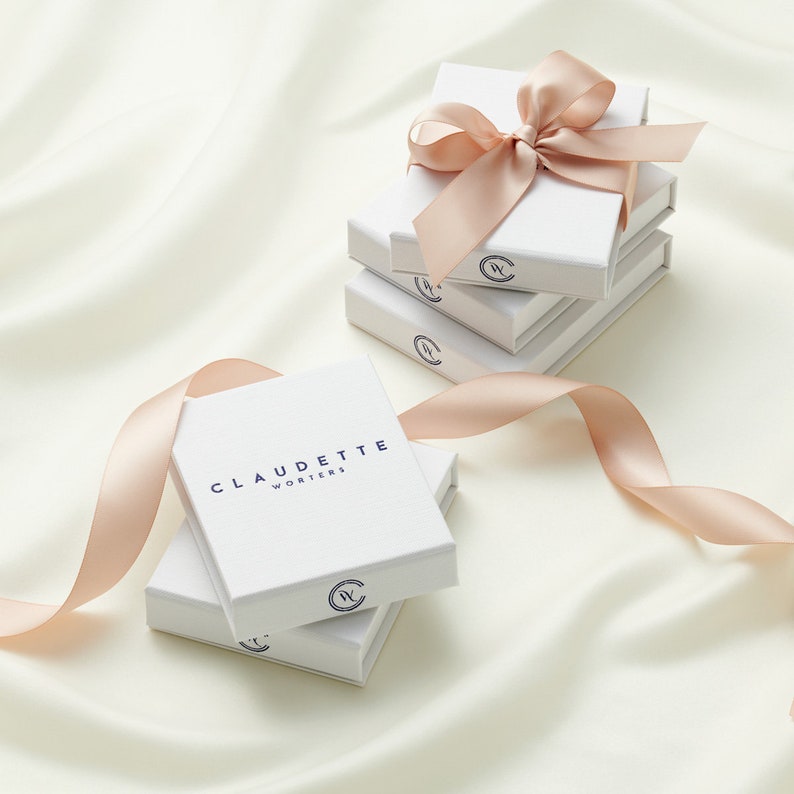 May include: A stack of three white gift boxes with a light pink ribbon on the top box. The top box has the text "CLAUDETTE WORKERS" printed on the front. The boxes are on a white surface.