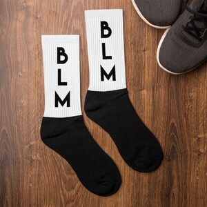 Può includere: Un paio di calzini bianchi con lettere nere che recitano "BLM" su ogni calzino. I calzini sono su una superficie di legno.
