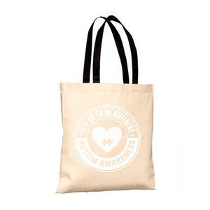 Può includere: Una borsa tote in tela beige con manici neri. La borsa ha un design circolare bianco con il testo "It's OK to be Different. Autism Awareness" e un cuore bianco con un pezzo di puzzle all'interno.
