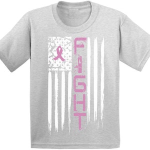 Pu&ograve; includere: Una maglietta grigia con un nastro rosa e la parola "FIGHT" stampata su un design di bandiera americana usurato.