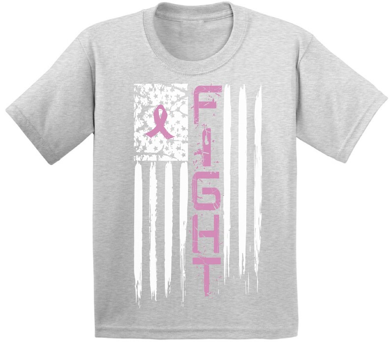 Pu&ograve; includere: Una maglietta grigia con un nastro rosa e la parola "FIGHT" stampata su un design di bandiera americana usurato.
