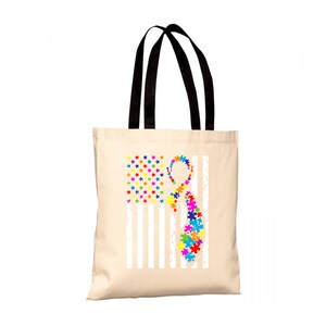 Può includere: Una borsa tote in tela bianca con manici neri. La borsa presenta un design di bandiera americana bianca e blu con un nastro colorato a forma di pezzo di puzzle.