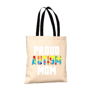 Può includere: Una borsa tote in tela beige con manici neri. La borsa ha la scritta "PROUD AUTISM MOM" stampata in bianco. La parola "AUTISM" è composta da pezzi di puzzle colorati.