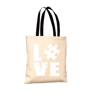 Può includere: Una borsa tote in tela beige con manici neri. La borsa ha una grafica bianca della parola "LOVE" con un pezzo di puzzle che sostituisce la "O". Il pezzo di puzzle ha un piccolo cuore al centro.