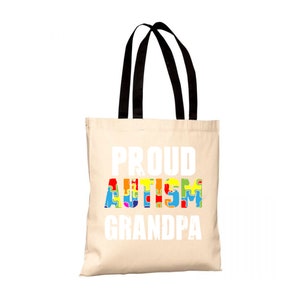Può includere: Una borsa tote in tela beige con manici neri. La borsa ha la scritta "PROUD AUTISM GRANDPA" stampata in bianco. La parola "AUTISM" è composta da pezzi di puzzle colorati.