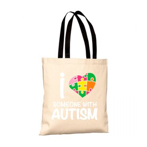 Può includere: Una borsa tote beige con manici neri. La borsa ha una grafica bianca di un cuore fatto di pezzi di puzzle e il testo "I ♥ SOMEONE WITH AUTISM".