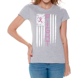 May include: A gray t-shirt with a white distressed American flag design. The flag has a pink ribbon in the upper left corner and the word "FIGHT" written vertically down the right side.