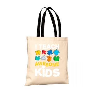 Può includere: Una borsa tote beige con manici neri. La borsa ha una grafica bianca che dice "Insegno bambini fantastici" con un design di pezzi di puzzle colorati.