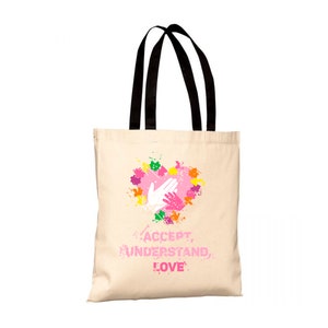Può includere: Una borsa tote color crema con manici neri. La borsa presenta un design a forma di cuore con schizzi di vernice colorati e due mani intrecciate. Le parole "ACCEPT, UNDERSTAND, LOVE" sono stampate in rosa sotto.