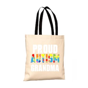 Può includere: Una borsa tote beige con manici neri. La borsa ha la scritta "PROUD AUTISM GRANDMA" stampata in bianco. La parola "AUTISM" è composta da pezzi di puzzle colorati.