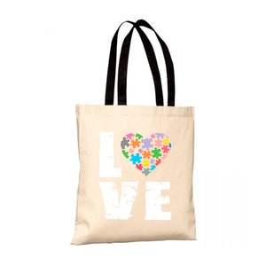 Può includere: Una borsa tote beige con manici neri. La borsa ha una grafica bianca della parola "LOVE" con un cuore fatto di pezzi di puzzle colorati al centro.
