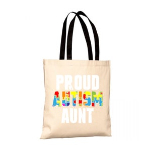 Può includere: Una borsa tote beige con manici neri. La borsa ha la scritta "PROUD AUTISM AUNT" stampata in bianco. La parola "AUTISM" è composta da pezzi di puzzle colorati.