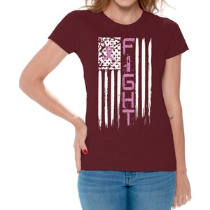 May include: A maroon t-shirt with a white and pink American flag design. The flag has the word "FIGHT" written vertically down the right side. The flag is distressed and has a pink ribbon in the top left corner.