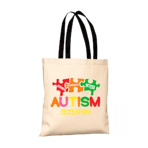 Può includere: Una borsa tote beige con manici neri. La borsa presenta una grafica colorata di pezzi di puzzle con le parole "Accetta", "Comprendi", "Ama" e "Consapevolezza dell'autismo".