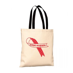Può includere: Una borsa tote in tela beige con manici neri. La borsa ha un nastro rosso con un motivo a pezzi di puzzle bianchi e il testo "AUTISM AWARENESS" stampato su di essa.