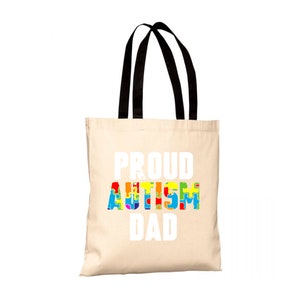 Può includere: Una borsa tote beige con manici neri. Le parole "PROUD AUTISM DAD" sono stampate in bianco sul davanti. La parola "AUTISM" è riempita con grafiche di pezzi di puzzle colorati.