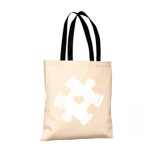 Può includere: Una borsa tote in tela beige con manici neri. La borsa presenta un pezzo di puzzle bianco invecchiato con un cuore al centro.