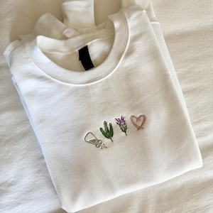 May include: White crewneck sweatshirt with embroidered designs. The embroidery includes a salt shaker, a sprig of rosemary, lavender, and a pink heart. The sweatshirt is made of a soft material.