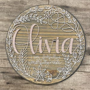 May include: A round wooden sign with a floral wreath design. The name "Olivia" is written in pink thread and the phrase "I am fearfully and wonderfully made" is written in white thread.