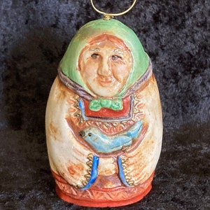 May include: A ceramic ornament of a woman wearing a green hooded cloak and holding a blue bird. The ornament has a gold loop for hanging.