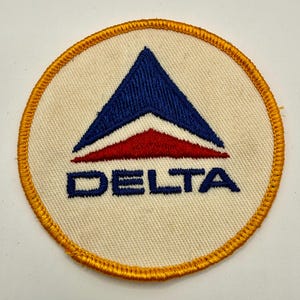 May include: A circular embroidered patch featuring an off-white fabric base with a gold-colored stitched border. The design includes a blue triangle above a red horizontal shape, with the word DELTA embroidered in blue capital letters below.