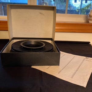 May include: A black slide carousel projector in its original box. The box is open, revealing the carousel with a circular design. A sheet of paper with printed text is next to the box. The box is black and beige.