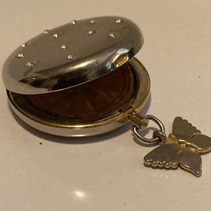 May include: A silver compact with a butterfly charm. The compact has a round, domed lid with a pattern of small, raised dots. The butterfly charm is gold and has a detailed design.