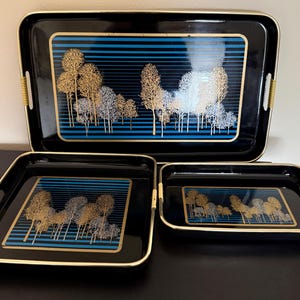 May include: A set of three black rectangular serving trays with gold-toned rims and integrated handles wrapped in light-colored material. Each tray features a central graphic of gold and silver trees against a background of horizontal blue stripes.