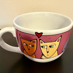 May include: A white ceramic mug featuring a wide pink band around the exterior. The band displays a graphic illustration of two cats, one orange and one yellow, positioned side by side beneath a small red heart. The mug has a single handle.