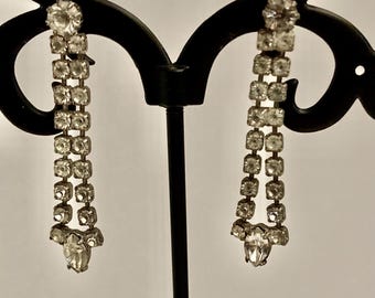 May include: A pair of silver tone dangle earrings with a row of clear rhinestones. The earrings are hanging from a black earring stand.
