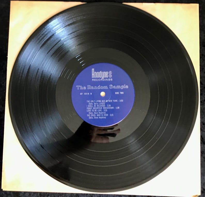 May include: A black vinyl record with a blue and white label. The label reads "Anodyne Recordings" and "The Random Sample." The track list includes "The Only Living Boy in New York" and "Foggy Mountain Breakdown." The record is in a tan paper sleeve.