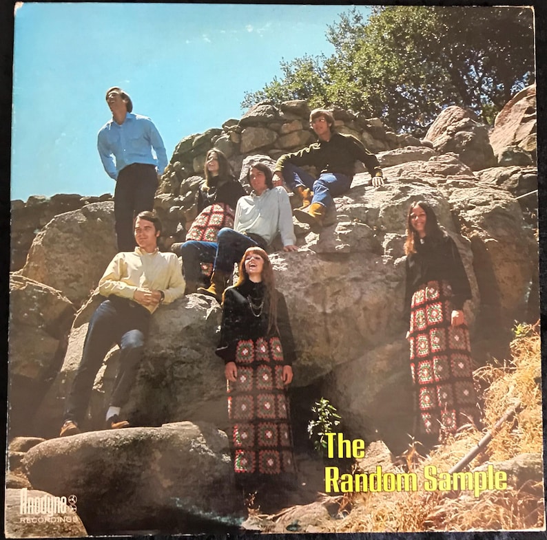 May include: Album cover for "The Random Sample" featuring a group of seven people posing on a rock formation. The album cover has a vintage aesthetic, with the band members dressed in 1960s or 1970s attire. The album title is in yellow text.