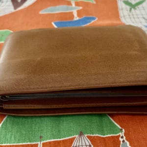 May include: A brown leather wallet, closed and slightly open, resting on a patterned orange and green surface. The wallet has a smooth, slightly glossy finish and appears to be well-worn.