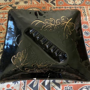 May include: Black square ashtray with gold leaf design. The ashtray has a built-in cigarette rest.