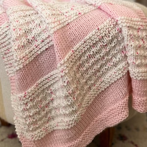 May include: A pink and white knitted baby blanket with a speckled pattern. The blanket is folded and draped over a chair.