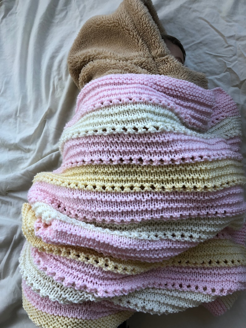 May include: A pink, white, and yellow striped knitted blanket with a brown fuzzy hooded sweater on top.