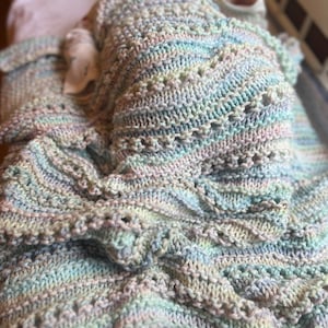 May include: A soft, multi-colored knitted baby blanket with a delicate, lacy pattern. The blanket is made of a light blue, pink, and white yarn.