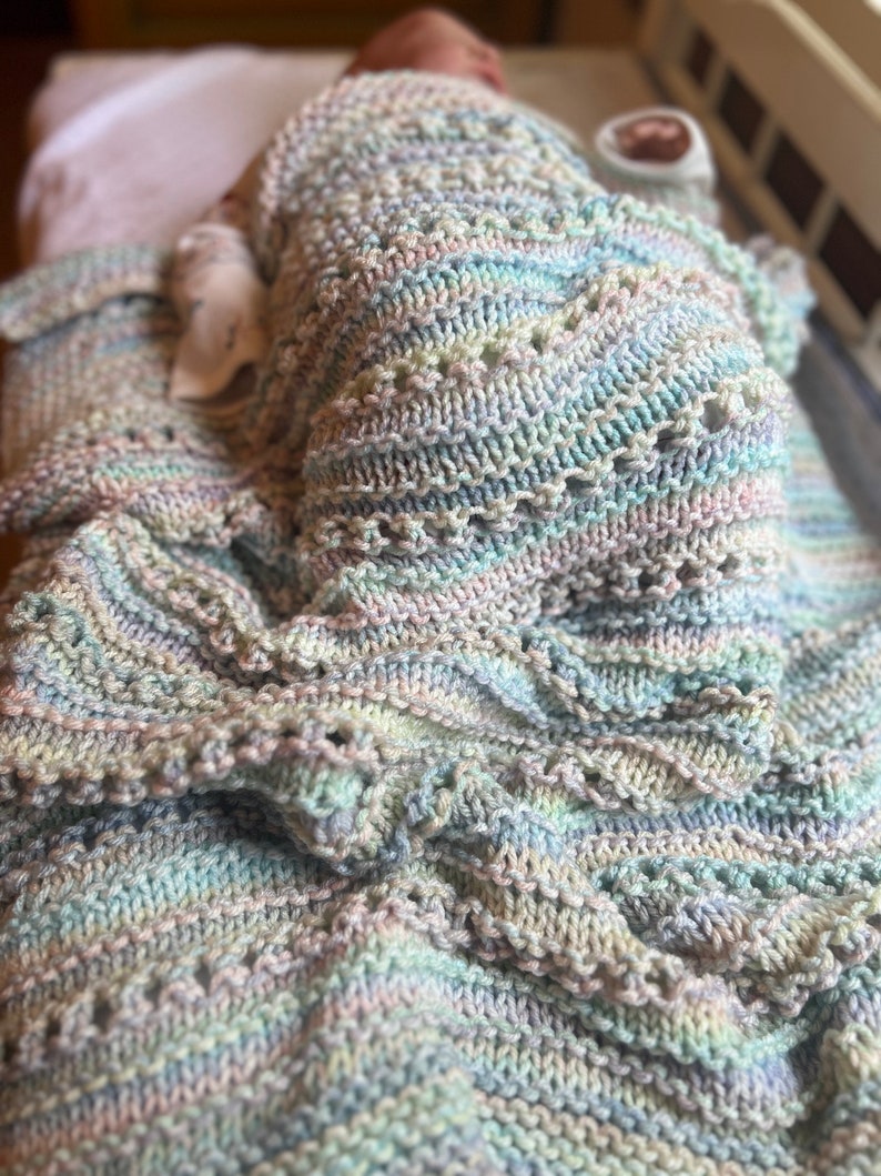 May include: A soft, multi-colored knitted baby blanket with a delicate, lacy pattern. The blanket is made of a light blue, pink, and white yarn.
