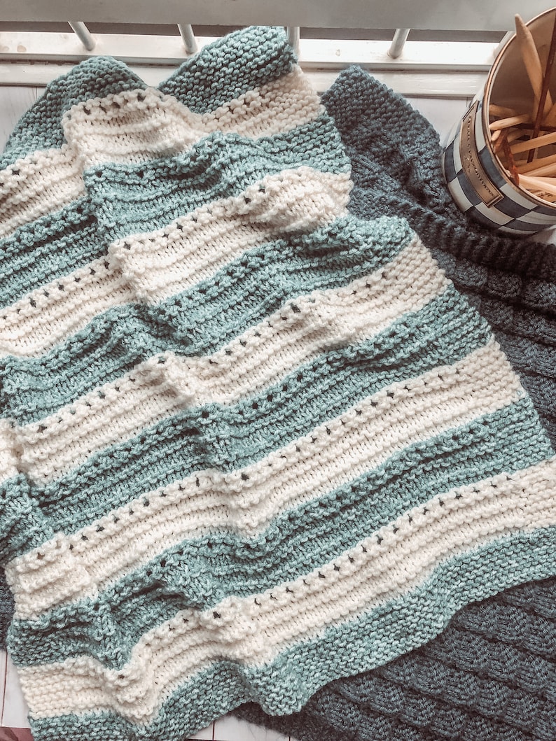 May include: A soft, striped knitted blanket in shades of teal and white. The blanket is folded and draped over a textured surface.