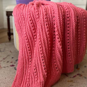 May include: A coral pink knitted blanket featuring a textured pattern of vertical columns and eyelet stitches. The blanket is draped over a surface, and the text "Beginner Friendly" with a yellow star icon is visible in the upper right corner.