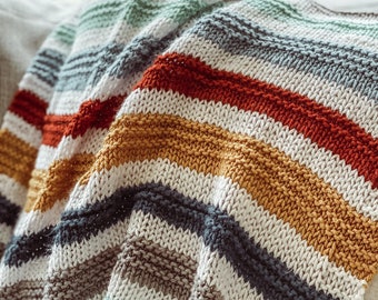 Könnte beinhalten: Eine gestreifte Strickdecke in den Farben Weiß, Grau, Blau, Grün, Gelb und Rot. Die Decke ist gefaltet und über ein Sofa gelegt.