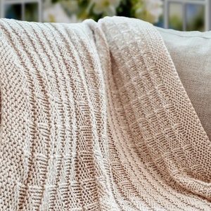 May include: A soft, beige knitted throw blanket with a textured pattern. The blanket is folded and draped over a couch.