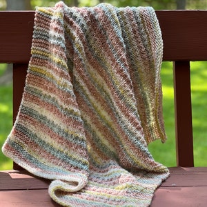 May include: A striped knitted blanket in shades of pink, green, white, and gray, draped over a wooden bench.
