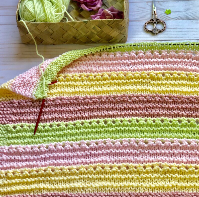 May include: A striped knitted blanket in shades of pink, yellow, and green. The blanket is being worked on with a red knitting needle.