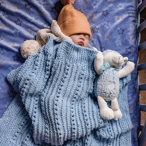 May include: A baby sleeping in a blue knitted blanket with a stuffed lamb toy. The baby is wearing a yellow knitted hat.