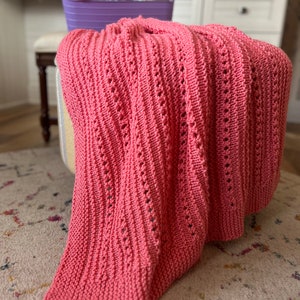 May include: A pink knitted throw blanket with a delicate open stitch pattern. The blanket is draped over a stool with a light brown and white striped fabric cover.