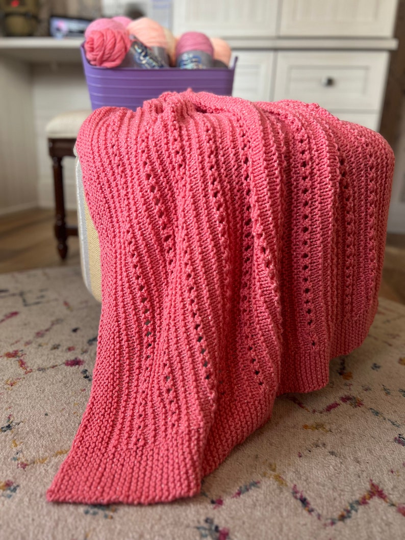 May include: A pink knitted throw blanket with a delicate open stitch pattern. The blanket is draped over a stool with a light brown and white striped fabric cover.
