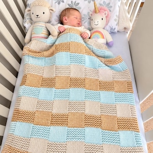 May include: A hand-knitted baby blanket featuring a checkered pattern in alternating squares of light blue, cream, and tan yarn. The blanket is draped over a sleeping infant in a crib, flanked by a plush sheep toy and a plush unicorn toy.